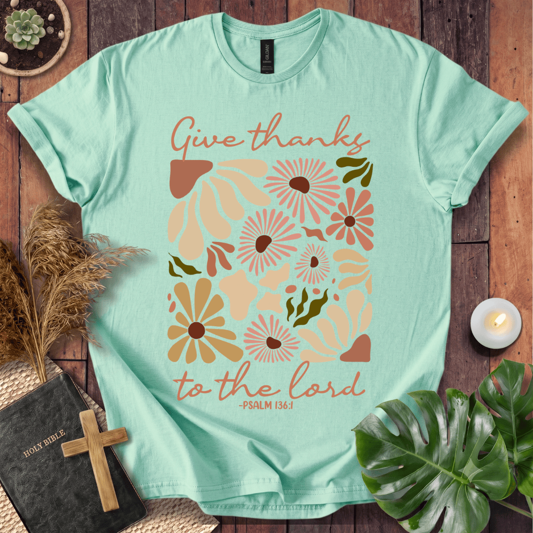Give Thanks to the Lord Psalm 136:1 white graphic t-shirt with retro boho rust and sage wildflowers on a wood background.