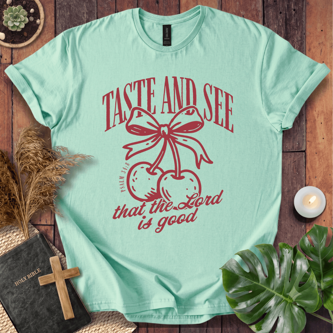 A mint green graphic t-shirt laid flat on a rustic wooden background, surrounded by decorative props including a Bible with a wooden cross, dried grass, a small candle, and a green plant leaf. The t-shirt features a red graphic of cherries tied with a bow and the text 'TASTE AND SEE that the Lord is good'.