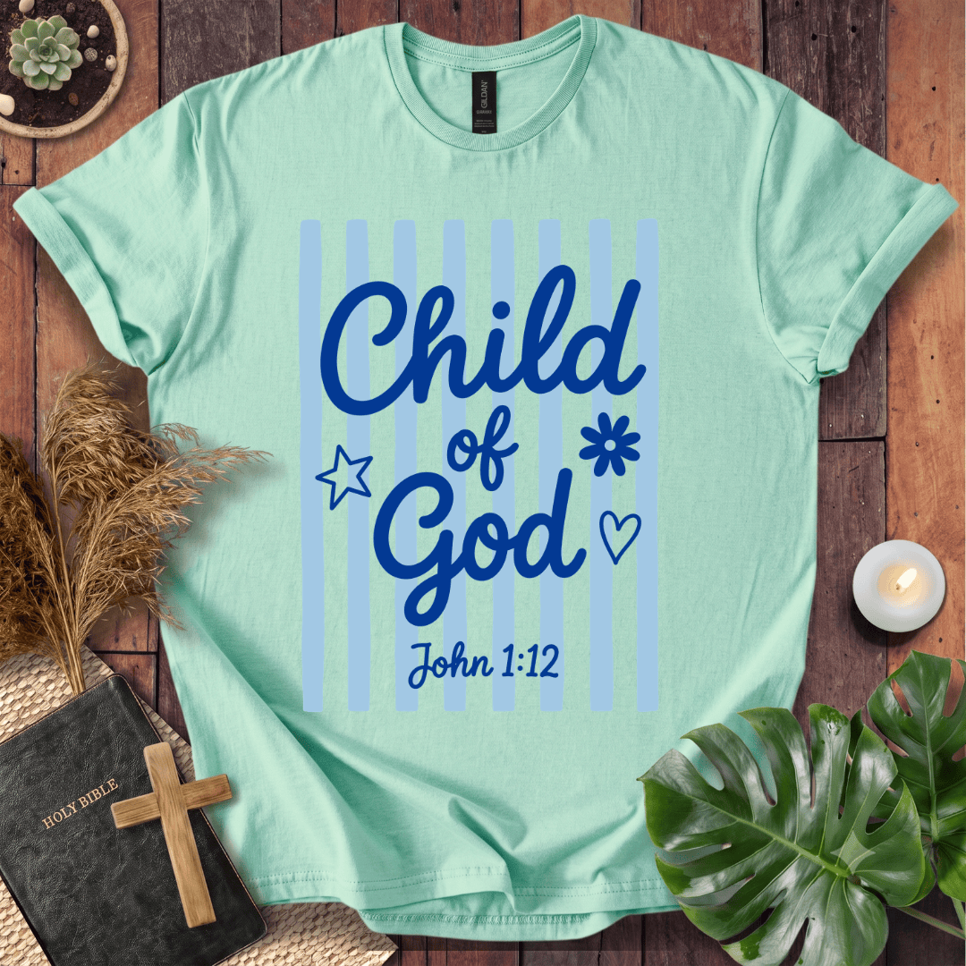 A mint green t-shirt with a blue graphic reading 'Child of God John 1:12' is displayed in a flat-lay arrangement on a wooden surface, surrounded by a bible, cross, pampas grass, a candle, and a monstera leaf.
