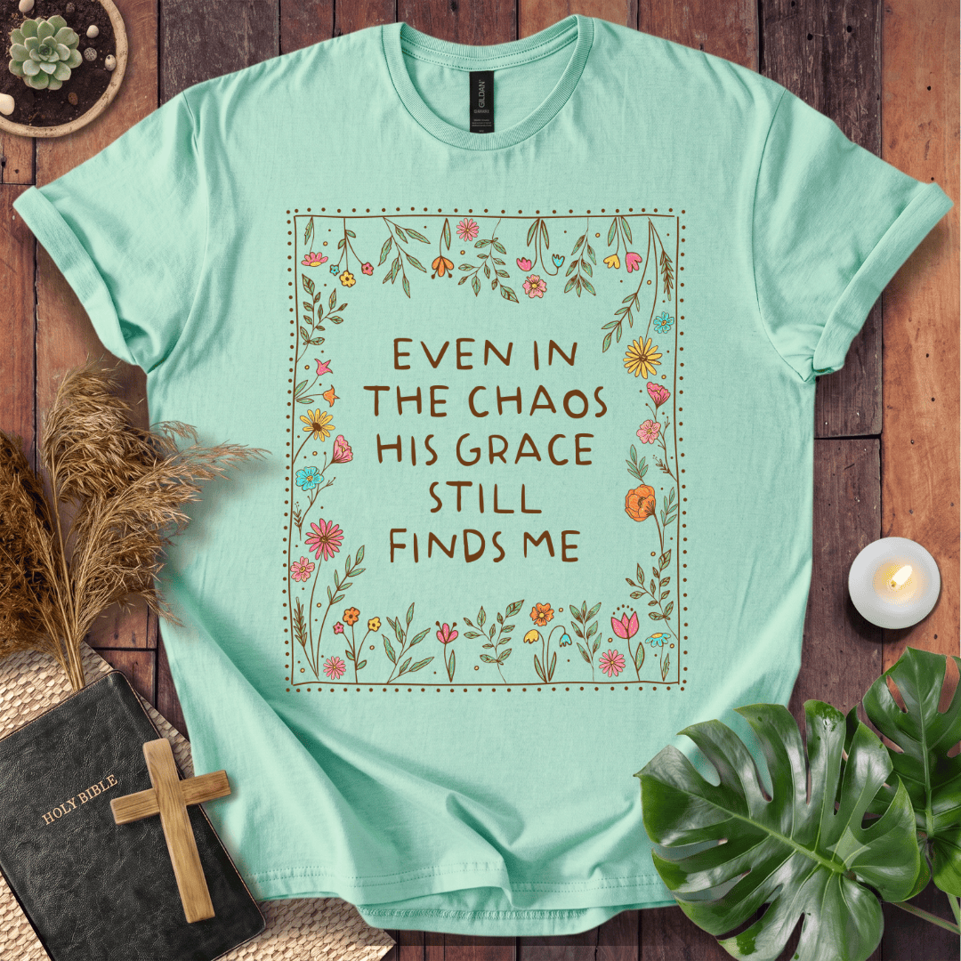 A mint green graphic t-shirt laid flat on a rustic wooden background, surrounded by decorative props including a bible with a wooden cross, dried grass, a small plant, a candle, and a monstera leaf. The t-shirt features a central floral border with the text 'EVEN IN THE CHAOS HIS GRACE STILL FINDS ME'.