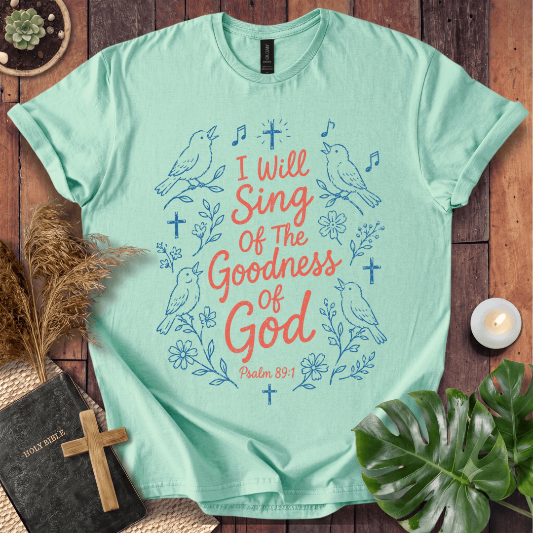 A mint green graphic t-shirt laid flat on a dark wood background, surrounded by decorative props including a Bible with a wooden cross, dried grass, a small candle, and a green leaf. The t-shirt features a blue line-art design of birds, flowers, and crosses with the text 'I Will Sing Of The Goodness Of God Psalm 89:1' in coral and blue lettering.