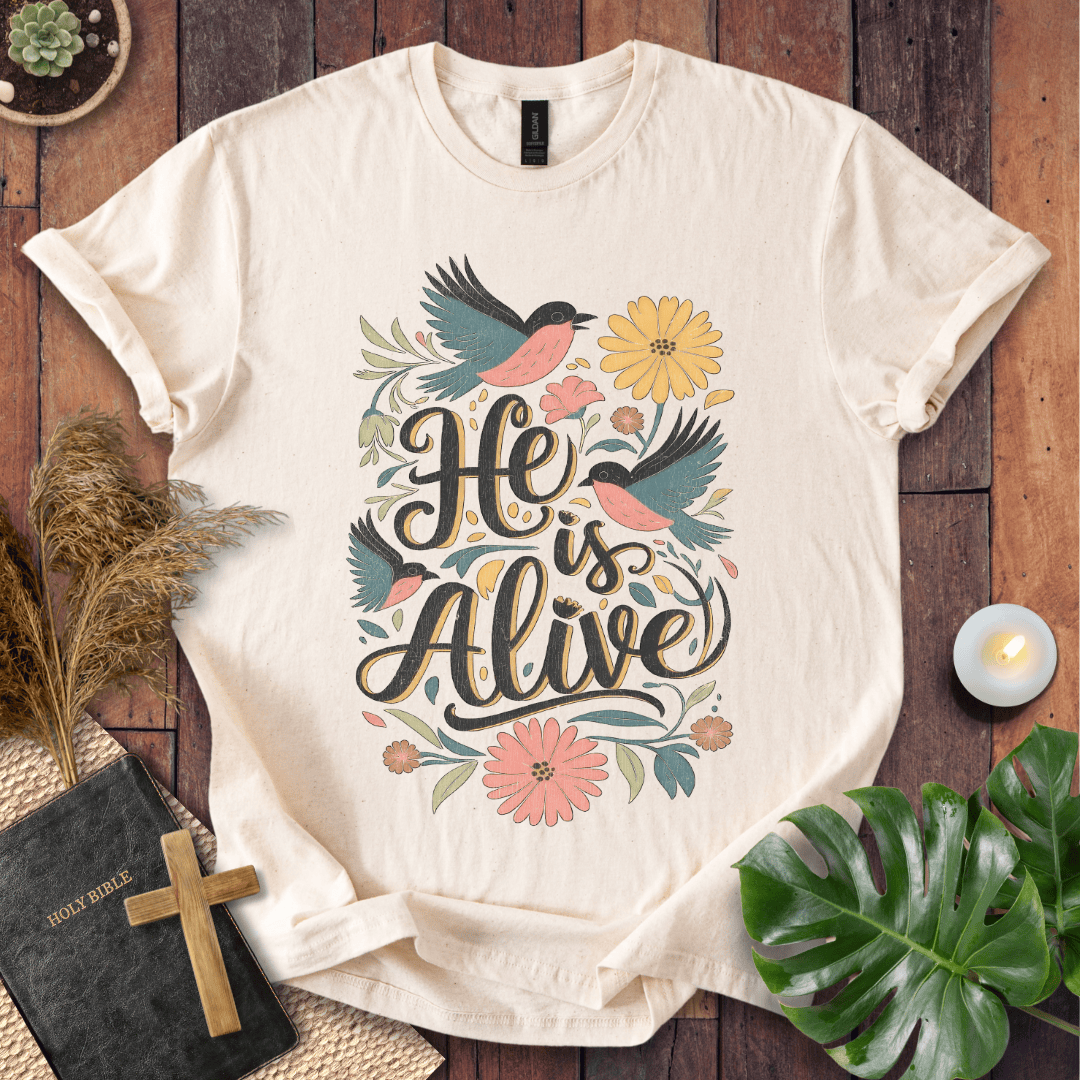 He Is Alive" Christian Easter t shirt for women with birds and flowers on a natural cream shirt, styled on a rustic wood background.
