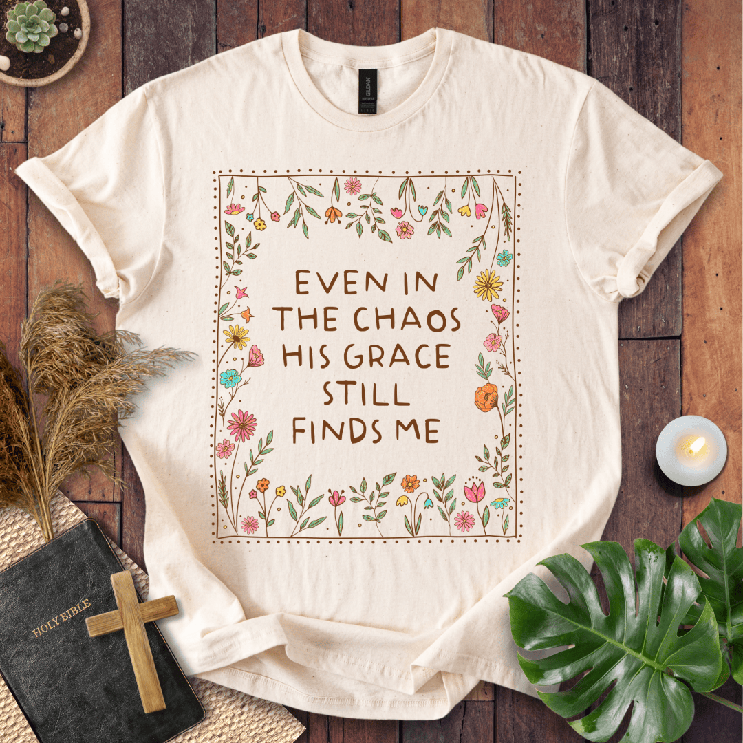 A cream-colored short-sleeve t-shirt with a floral graphic and the text 'EVEN IN THE CHAOS HIS GRACE STILL FINDS ME' printed on the front. The shirt is styled in a flat-lay arrangement on a dark wood background with a bible, wooden cross, pampas grass, a lit candle, and a green monstera leaf.