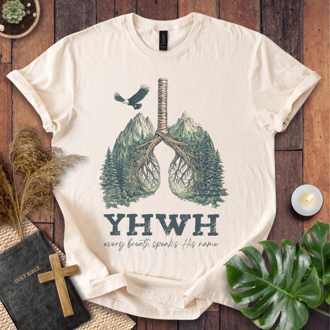 Christian t-shirt with YHWH design, tree-lung illustration, mountains, and “Every Breath Speaks His Name” text, laid flat with Bible and plants.