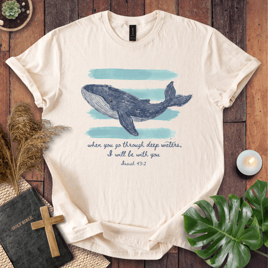 A cream-colored short-sleeve t-shirt with a graphic of a blue-gray whale on a light blue watercolor background, with the text 'when you go through deep waters, I will be with you Jesus 48:2' printed below. The shirt is displayed in a flat-lay arrangement on a dark wood background with decorative props including a bible, wooden cross, dried grass, a small candle, and a green monstera leaf.