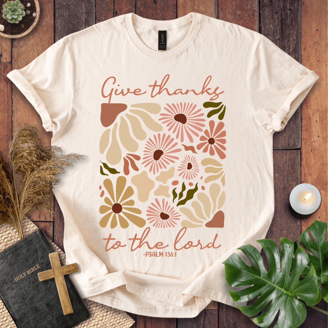 Give Thanks to the Lord Psalm 136:1 white graphic t-shirt with retro boho rust and sage wildflowers on a wood background.