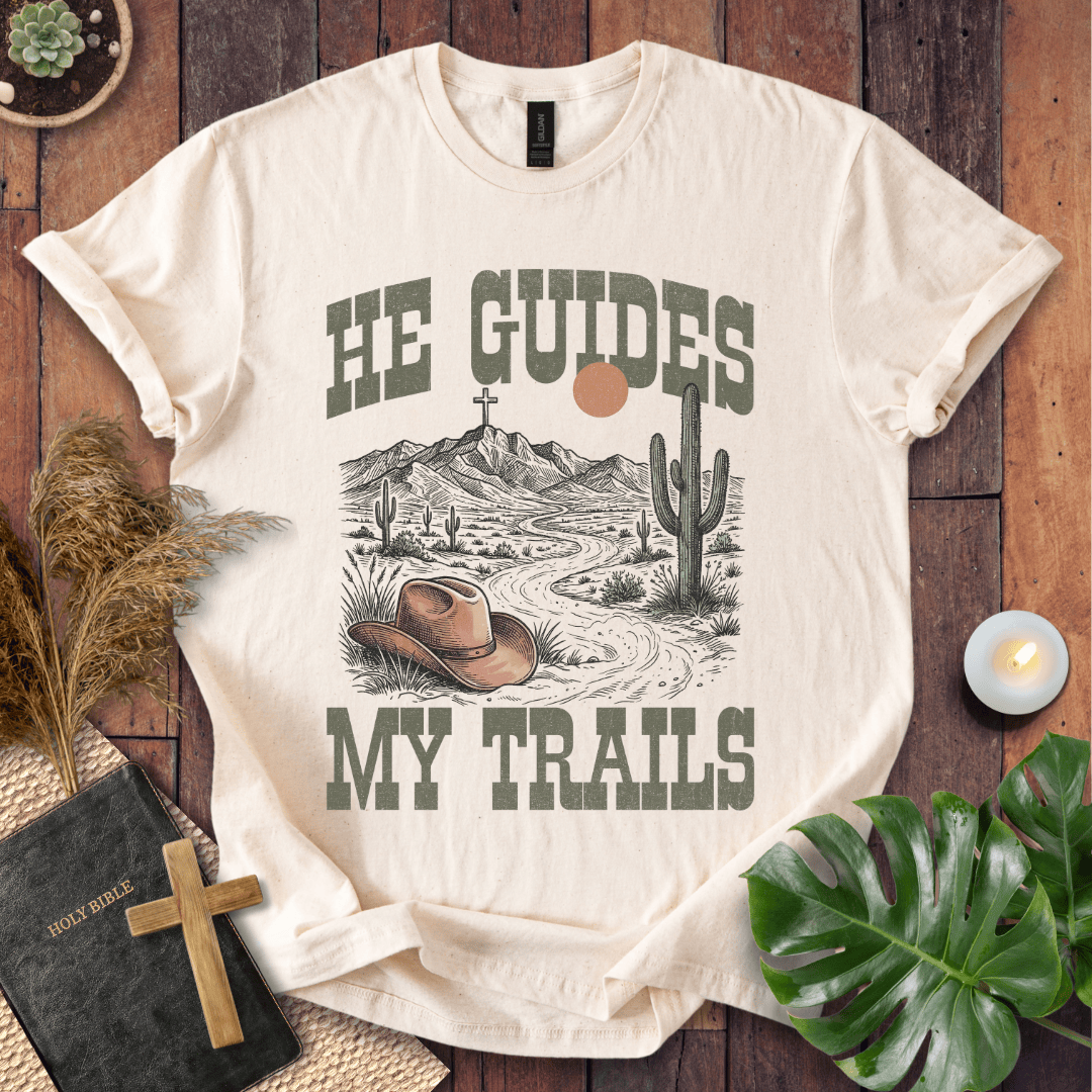 A Christian t-shirt with a desert landscape, cowboy hat, and cross, reading He Guides My Trails.