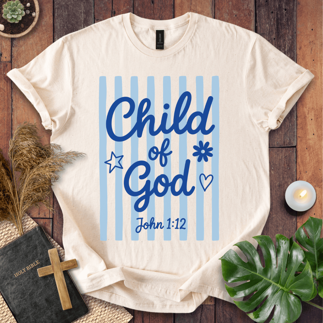 A cream-colored t-shirt with a blue graphic reading 'Child of God John 1:12' on a background of light blue vertical stripes, styled in a flat-lay on a wooden surface with a bible, cross, pampas grass, and a lit candle.