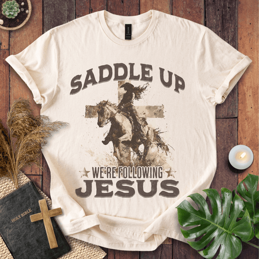 t-shirt featuring a cowgirl riding a horse in front of a cross with the text Saddle Up We're Following Jesus.