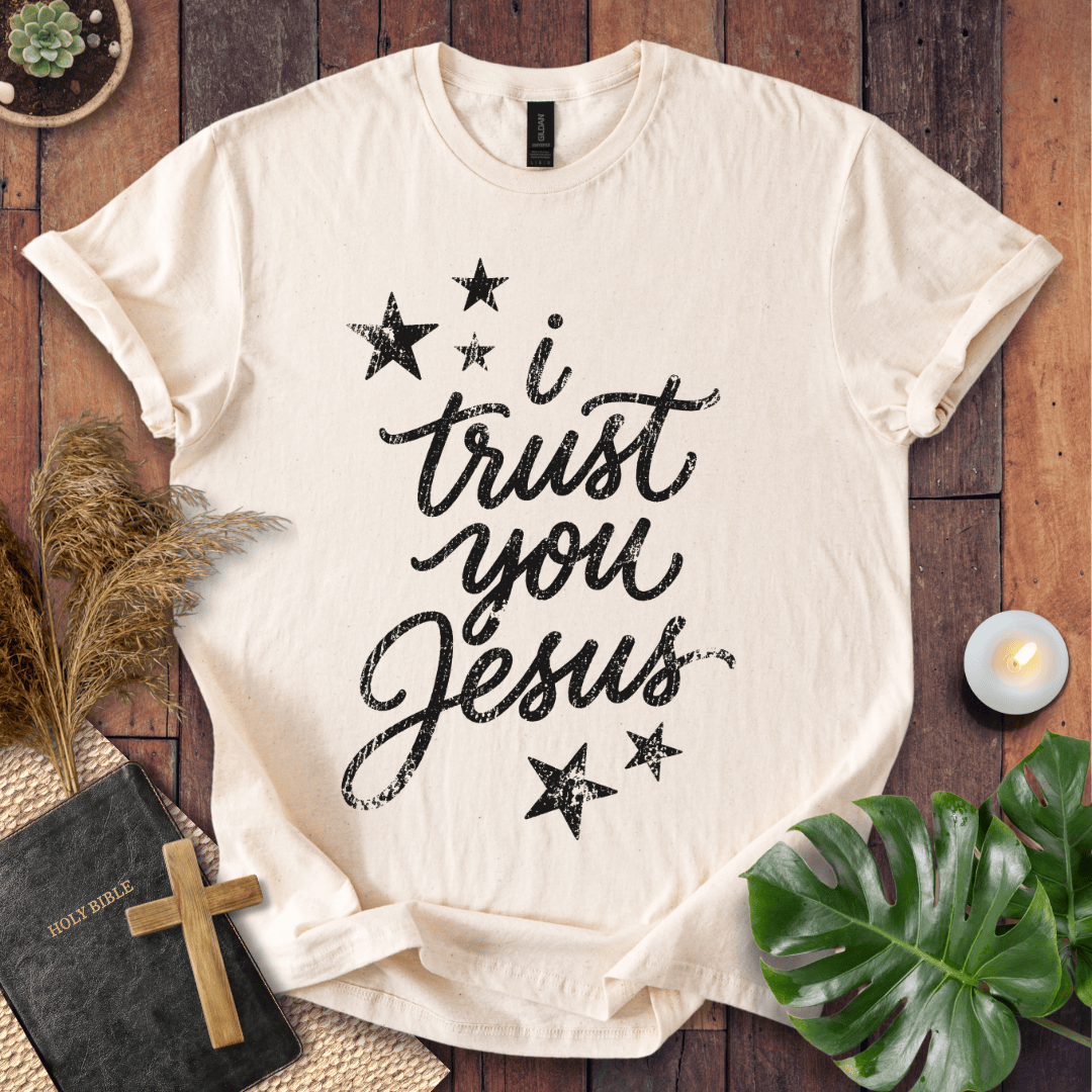 A cream-colored short-sleeve t-shirt with black text reading 'i trust you Jesus' and black stars, laid flat on a dark wood background with decorative props including a book, cross, pampas grass, candle, and green leaves.