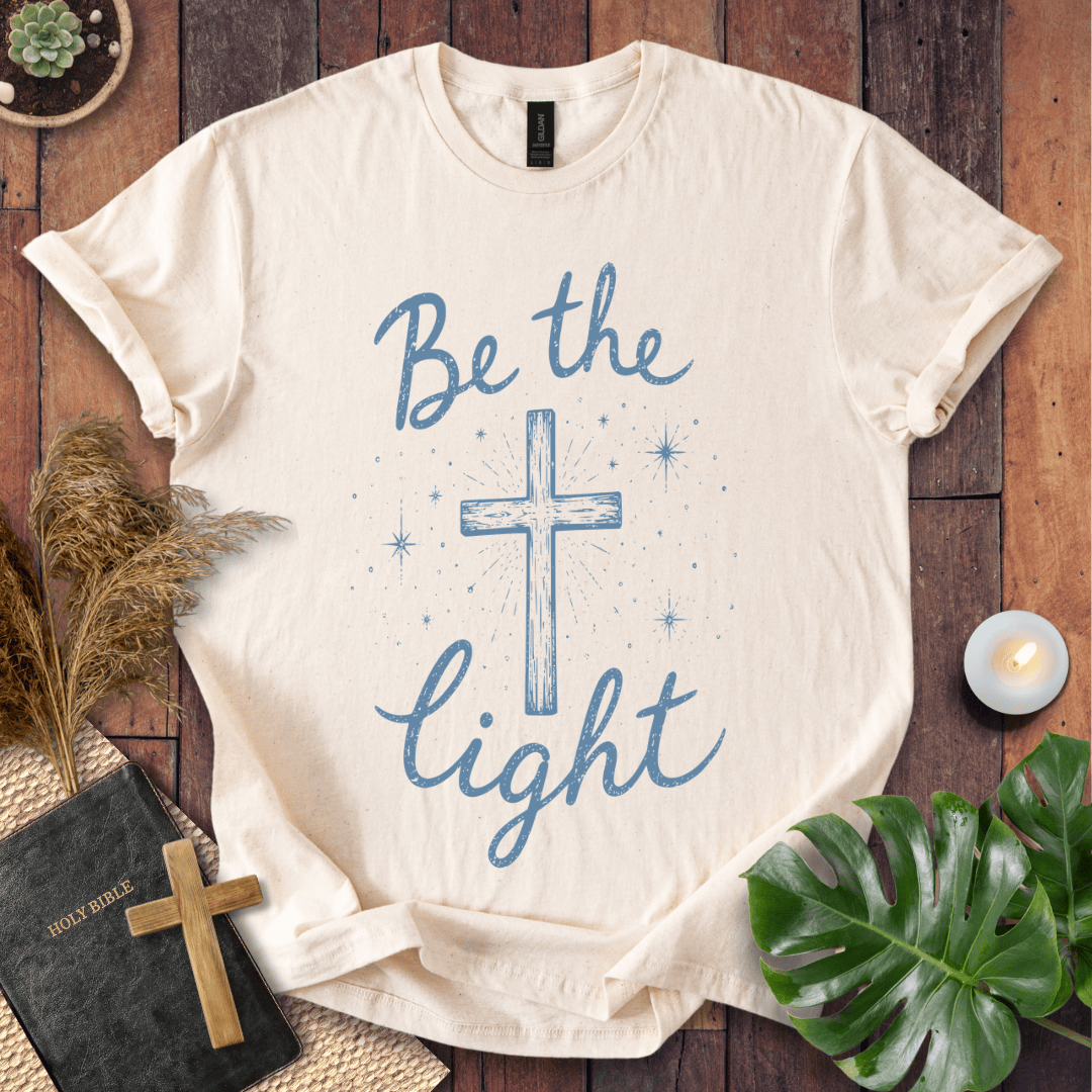 Christian t shirt for women with blue "Be the Light" text, a central cross, and starry accents on a wooden background.
