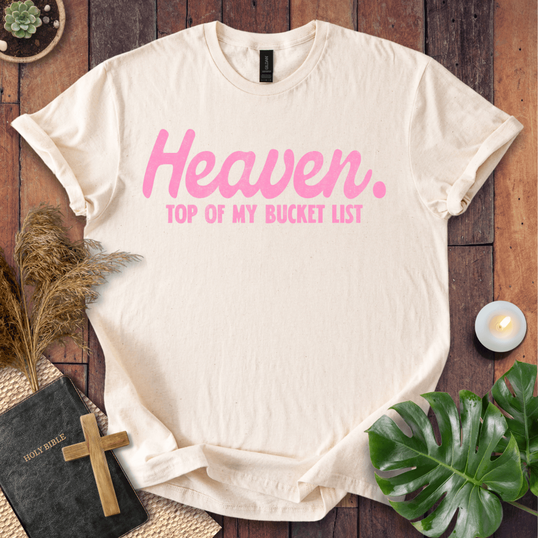 A cream-colored short-sleeve t-shirt with pink text reading 'Heaven. TOP OF MY BUCKET LIST' laid flat on a dark wood background with decorative props including a book, cross, candle, pampas grass, and green leaves.