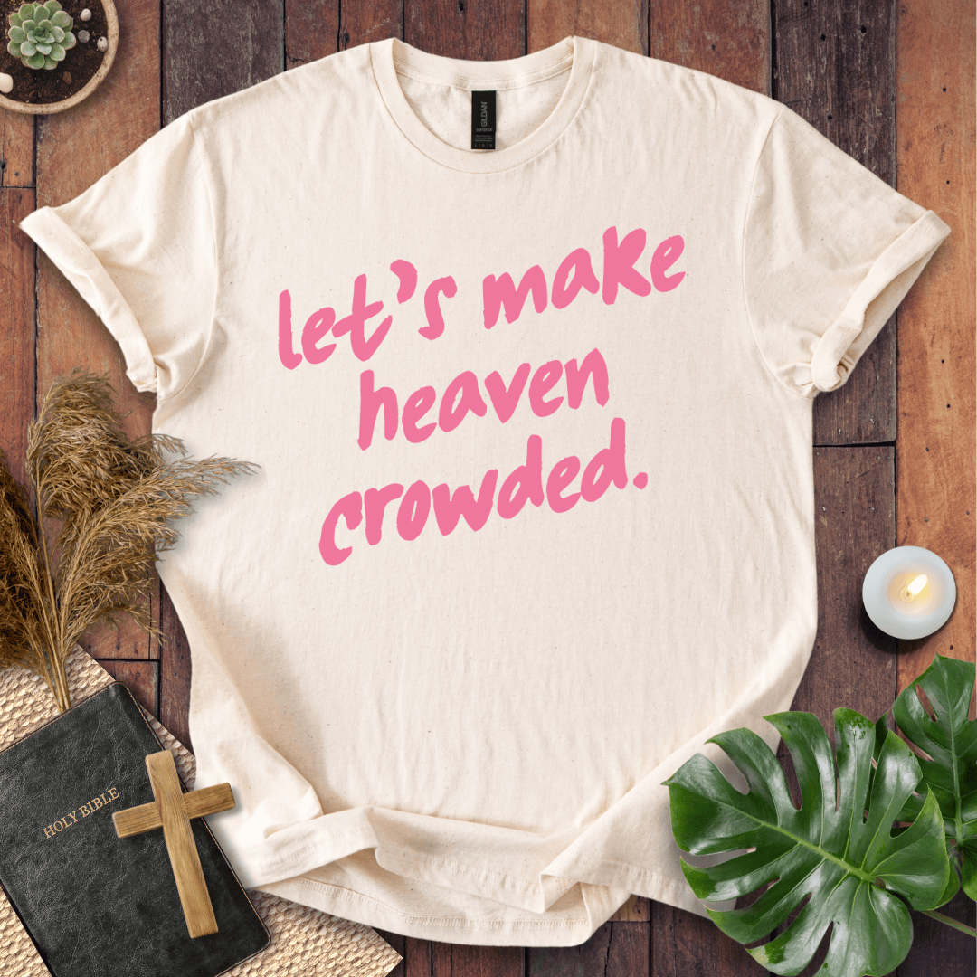 A cream-colored short-sleeve t-shirt with pink text reading 'let's make heaven crowded.' laid flat on a dark wood background with decorative props including a Bible, cross, candle, dried grass, and green leaves.