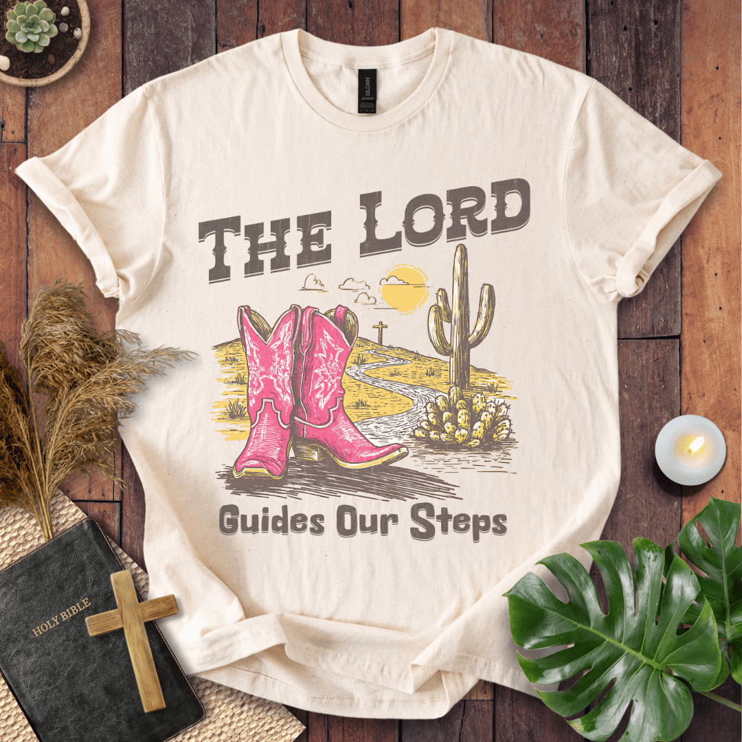 t-shirt with pink cowboy boots and "The Lord Guides Our Steps" text in a desert landscape with a cross and cactus.