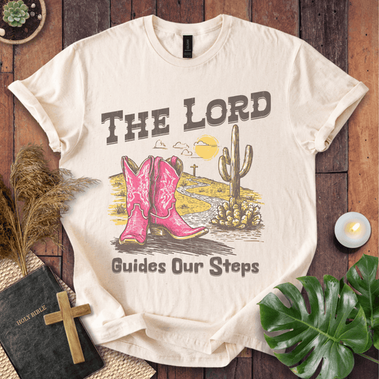 t-shirt with pink cowboy boots and "The Lord Guides Our Steps" text in a desert landscape with a cross and cactus.