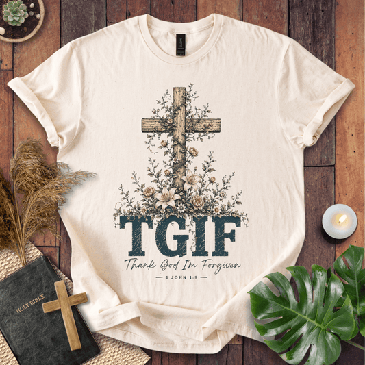 A cream-colored short-sleeve t-shirt with a rustic graphic print featuring a wooden cross, floral details, and the text 'TGIF Thank God I'm Forgiven JON 1:9'. The t-shirt is displayed in a styled flat-lay on a dark wood background, surrounded by a Bible, a small wooden cross, dried grass, a candle, and green leaves.