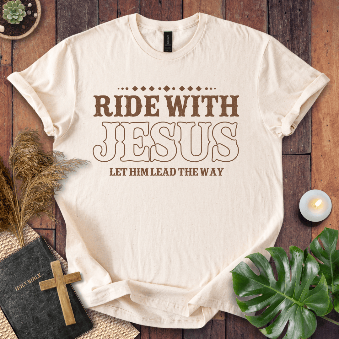 Ride With Jesus Western graphic tee on a wooden background with a Bible, cross, and green leaves.