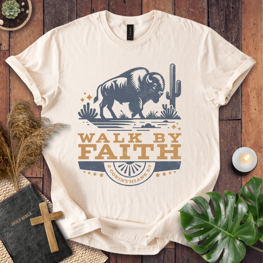 Beige t-shirt with a graphic of a bison and 'Walk by Faith' text on a wooden surface.