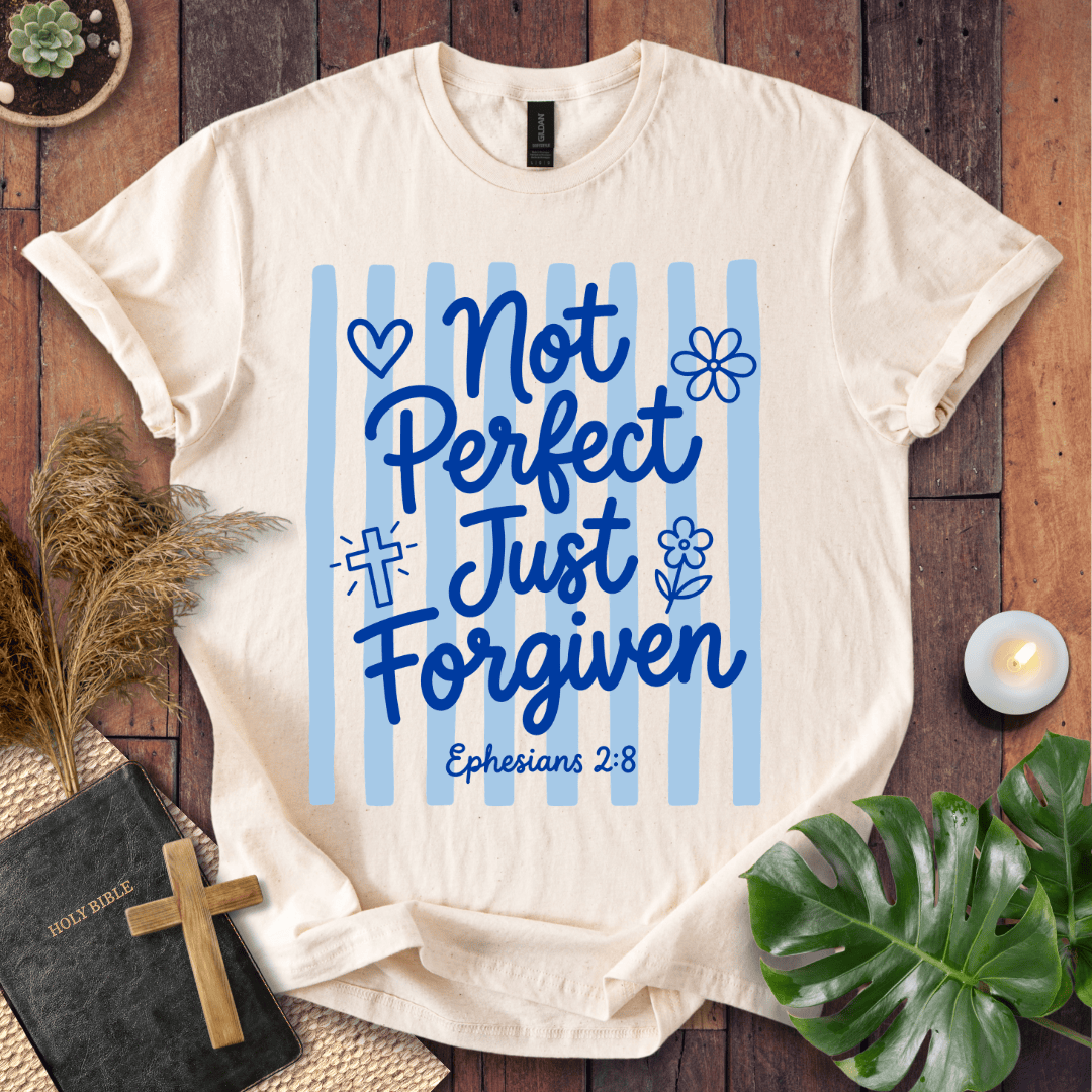 A cream-colored t-shirt with a blue graphic reading 'Not Perfect Just Forgiven Ephesians 2:8' on light blue vertical stripes, styled in a flat-lay on a wooden surface with a Bible, cross, pampas grass, and green leaves.