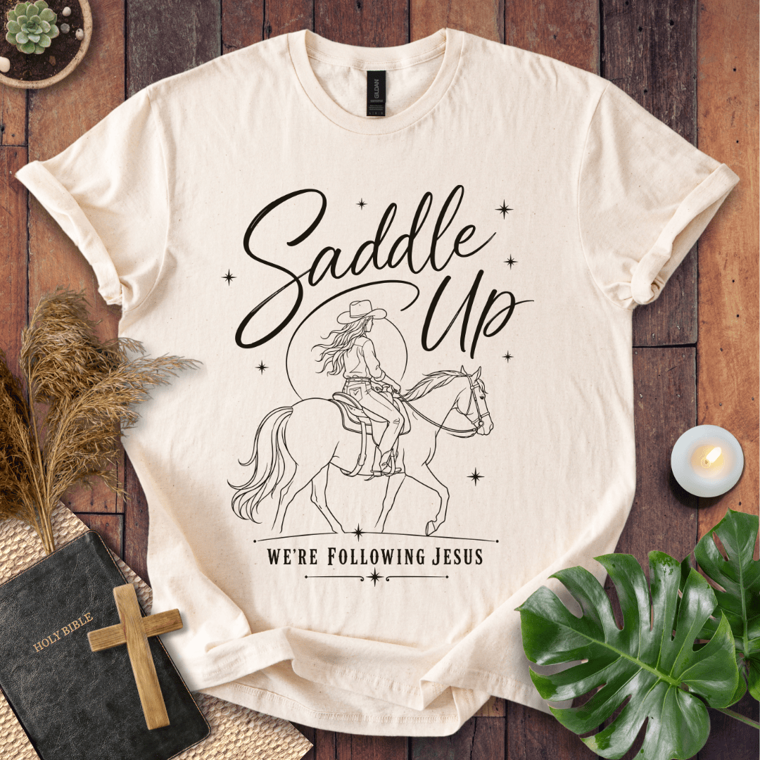 "Saddle Up, We're Following Jesus" shirt with a cowgirl riding a horse under a moonlit sky, displayed on a wooden background.