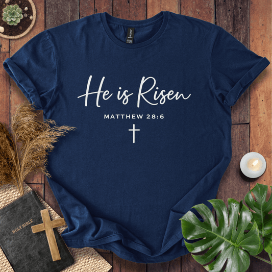 Easter Christian t-shirt with white "He is Risen" script and cross, styled on wood with a Bible and wooden cross.