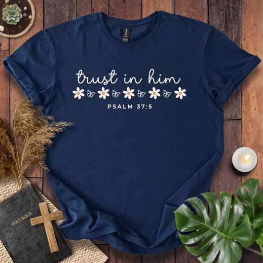 A navy blue short-sleeve T-shirt laid flat on a wooden surface, printed with the phrase 'trust in him' in white script, small white flowers, and 'PSALM 37:5' below. The shirt is styled with a book, cross, dried grass, a small candle, and green leaves around it.