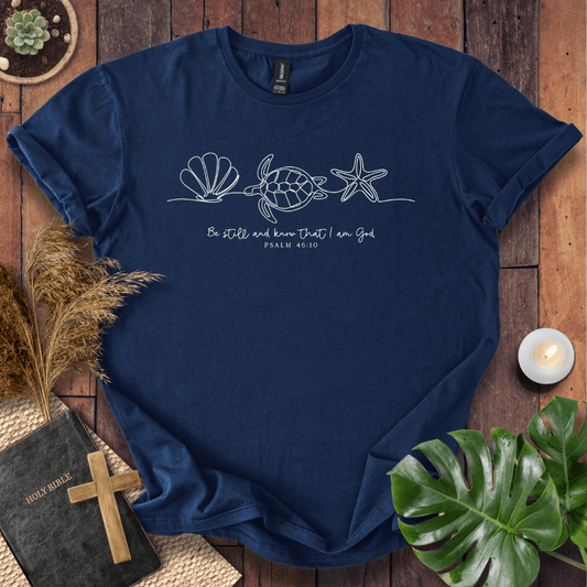 A navy blue short-sleeve t-shirt with a white line art graphic of a seashell, sea turtle, and starfish, with the text 'Be still and know that I am God PSALM 46:10' printed on the chest. The shirt is presented in a styled flat-lay on a wooden surface with a Bible, wooden cross, pampas grass, a small candle, and a green monstera leaf.