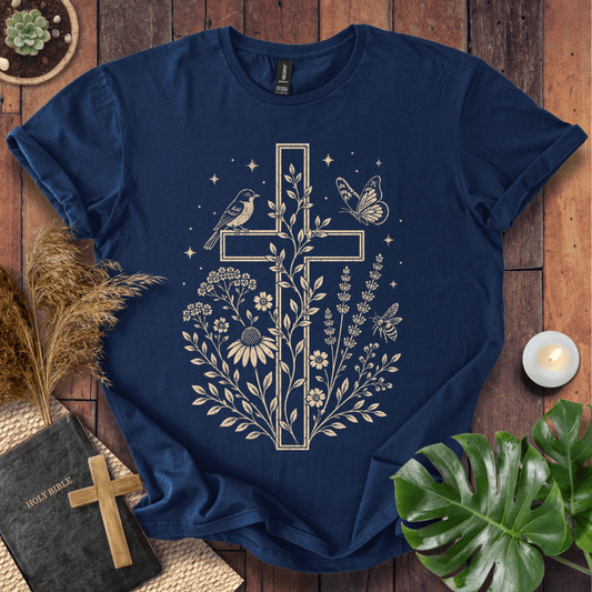 A navy blue short-sleeve t-shirt with a cream-colored graphic of a cross surrounded by wildflowers, a bird, and a butterfly, laid flat on a rustic wooden surface with decorative props including a Bible, dried grass, a candle, and a green leaf.