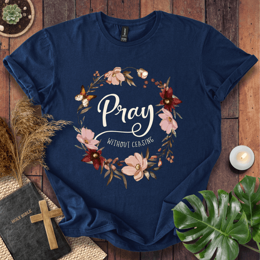 "Pray Without Ceasing" text inside a floral wreath with a butterfly on a shirt next to a Bible and cross.