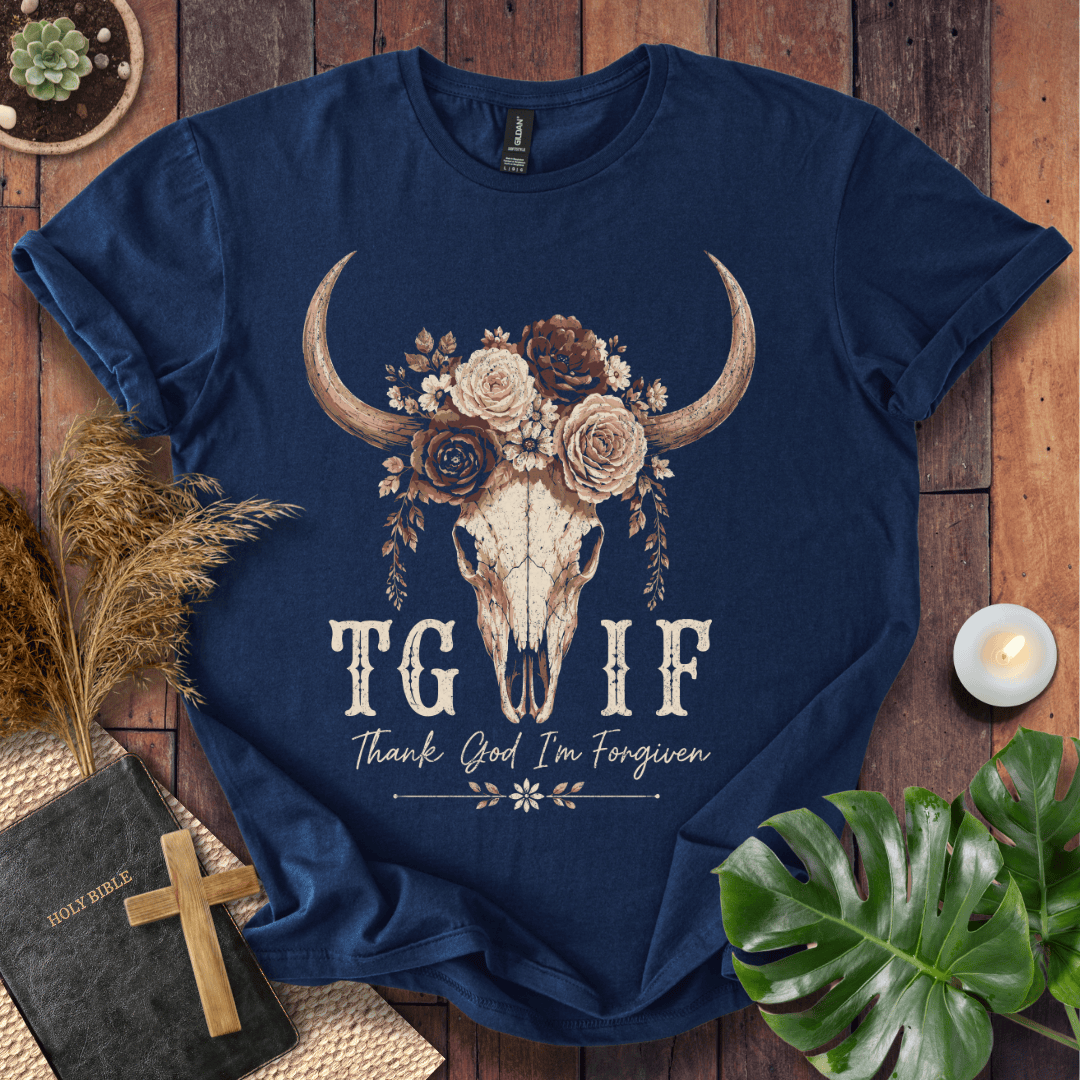 A navy blue graphic t-shirt laid flat on a rustic wooden background with decorative props including a book, cross, dried grass, monstera leaf, and candle. The shirt features a large floral and horned skull graphic above the text 'TG IF' and 'Thank God I'm Forgiven'.