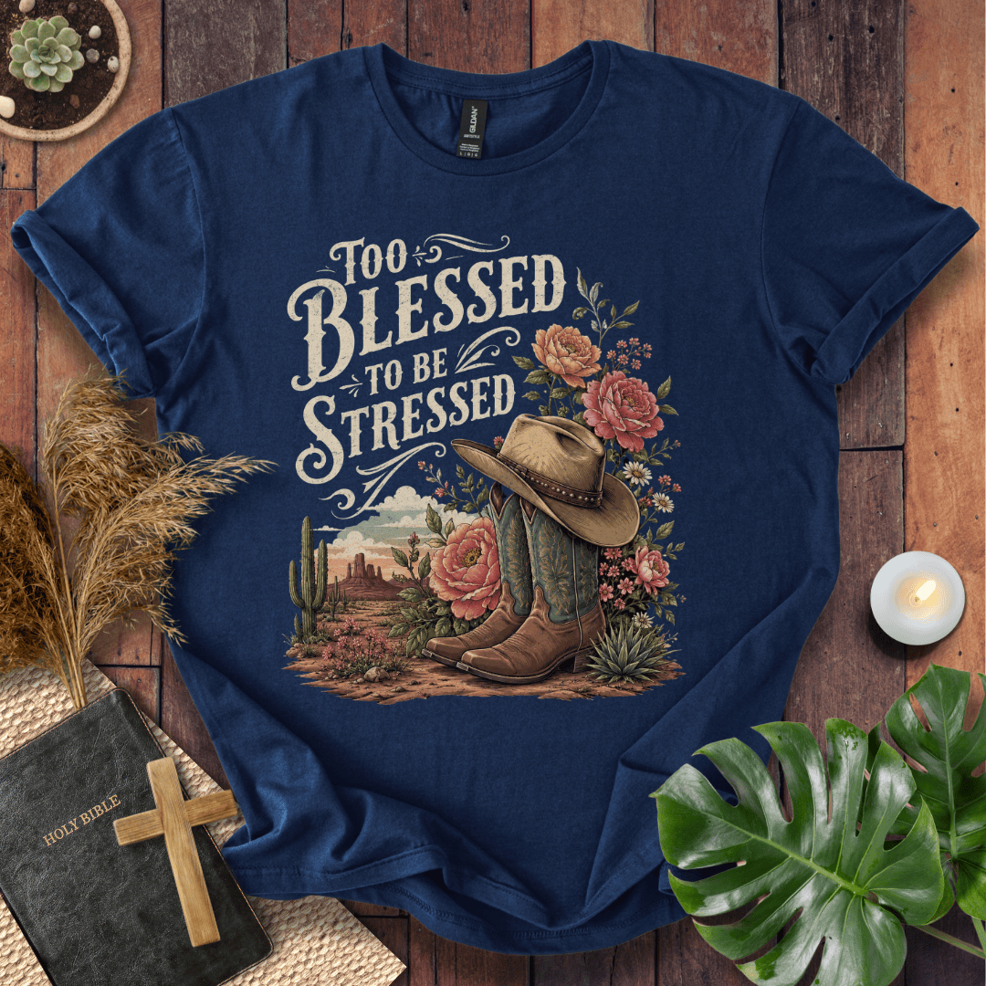 A navy blue graphic t-shirt laid flat on a rustic wooden surface, surrounded by decorative props including a Bible with a cross, dried grass, a small candle, and green leaves. The t-shirt features a western-themed graphic with the text 'Too Blessed to be Stressed' above a cowboy hat, boots, cactus, and flowers.