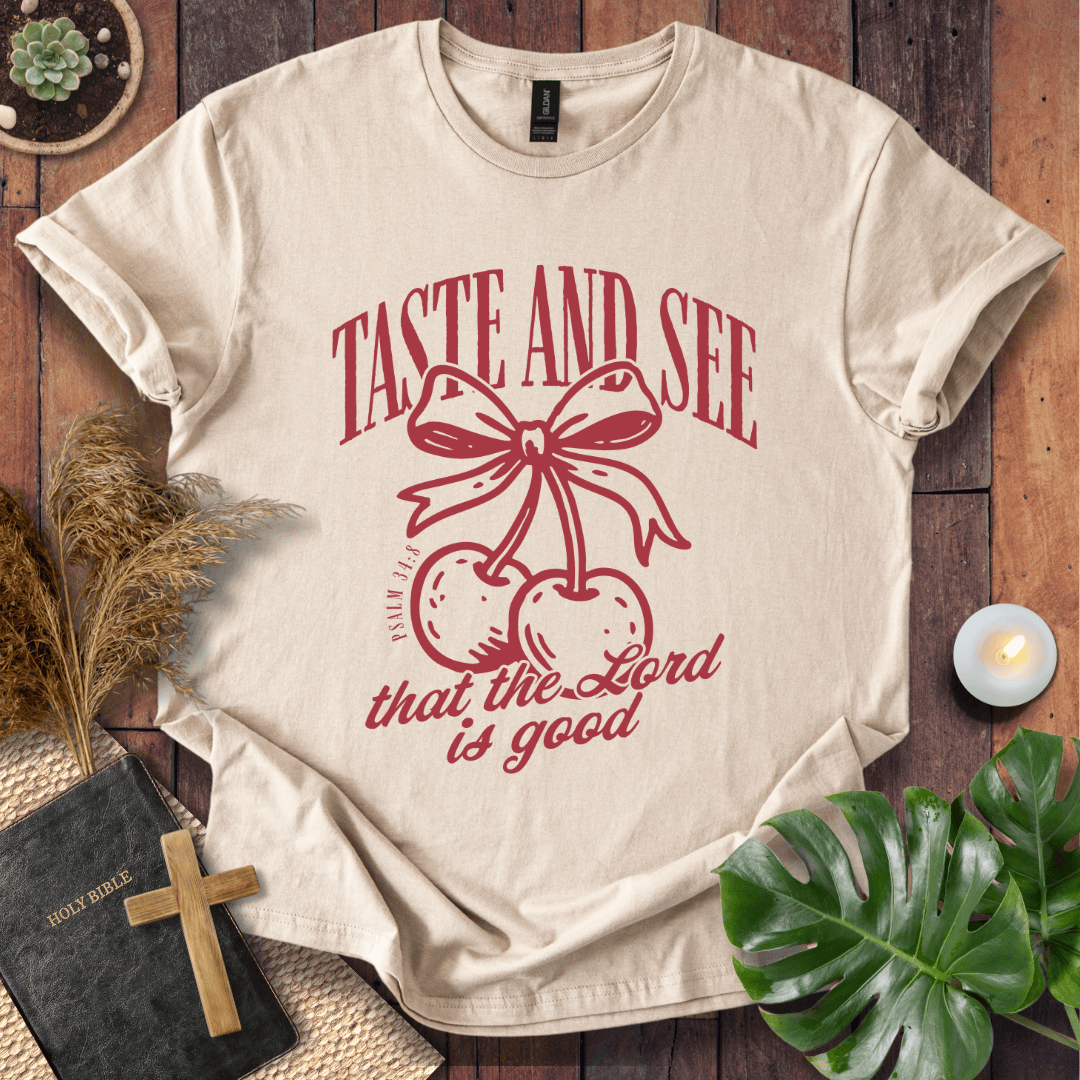 A beige short-sleeve graphic T-shirt laid flat on a rustic wooden surface, surrounded by decorative props including a book with a wooden cross, dried grass, a small candle, and green leaves. The shirt has a red printed design featuring cherries tied with a bow and the text 'TASTE AND SEE that the Lord is good'.