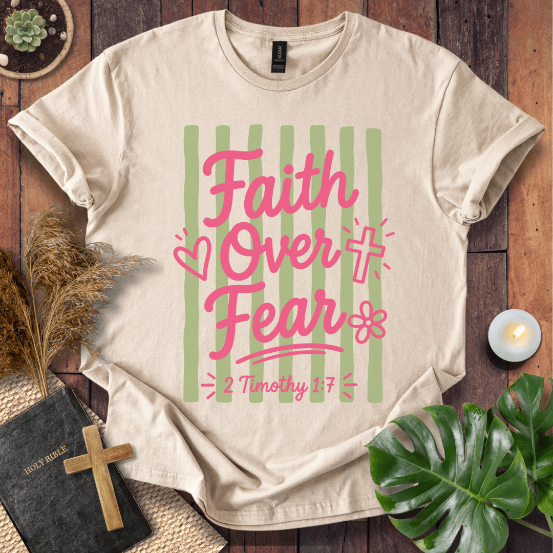 A beige short-sleeve graphic T-shirt laid flat on a rustic wood surface, featuring the phrase 'Faith Over Fear' in pink script over vertical light green stripes, with small heart, cross, and flower motifs. The shirt is styled with a Bible, wooden cross, dried grass, plant leaves, and a small candle.