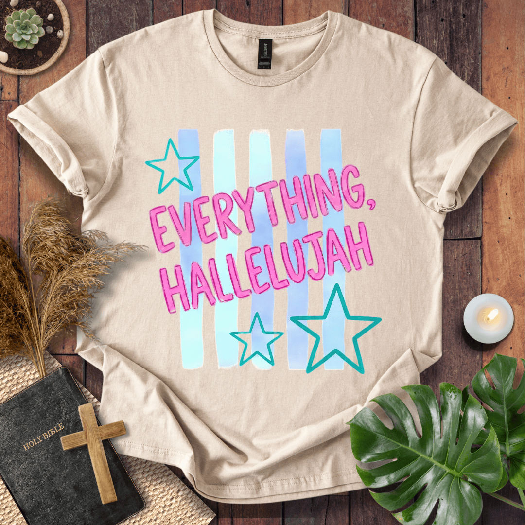 A beige graphic t-shirt laid flat on a rustic wooden surface, surrounded by decorative props including a small cross, a book, dried grass, a plant leaf, and a candle. The t-shirt features the text 'EVERYTHING, HALLELUJAH' in pink letters over light blue vertical stripes and teal star outlines.