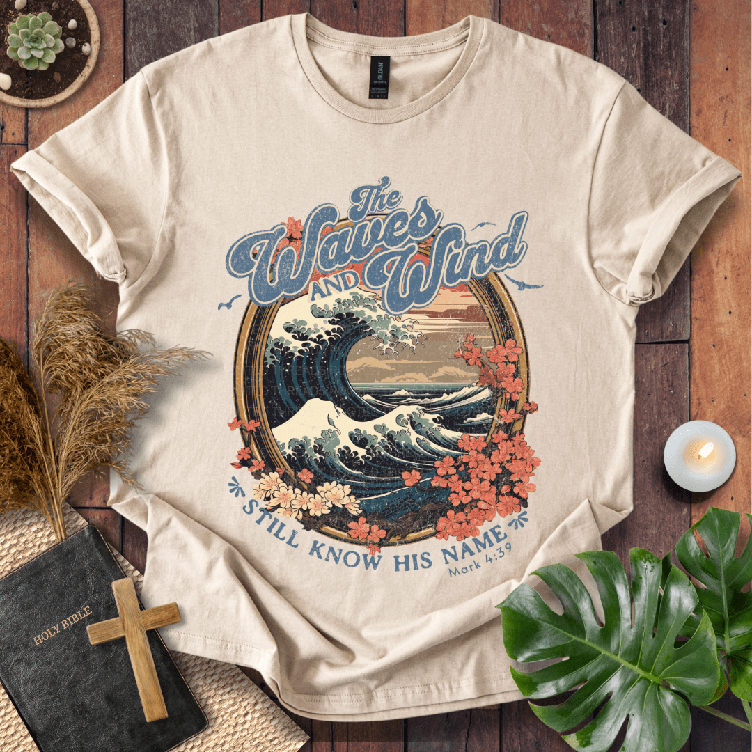 The Waves and Wind Still Know His Name Christian t-shirt in sand—vintage wave, sunset, and floral frame, Mark 4:39.
