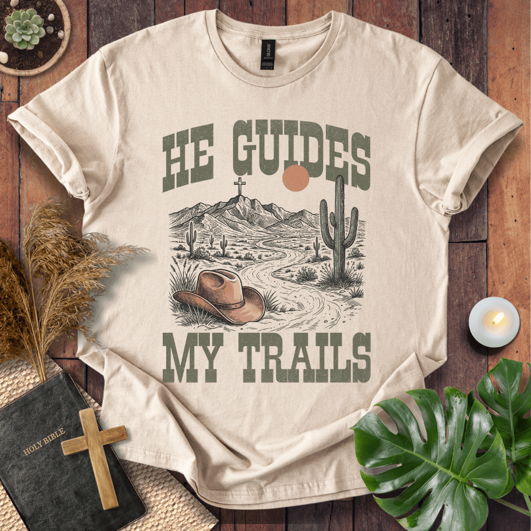 A Christian t-shirt with a desert landscape, cowboy hat, and cross, reading He Guides My Trails.