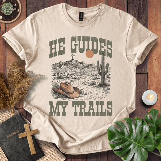 A Christian t-shirt with a desert landscape, cowboy hat, and cross, reading He Guides My Trails.
