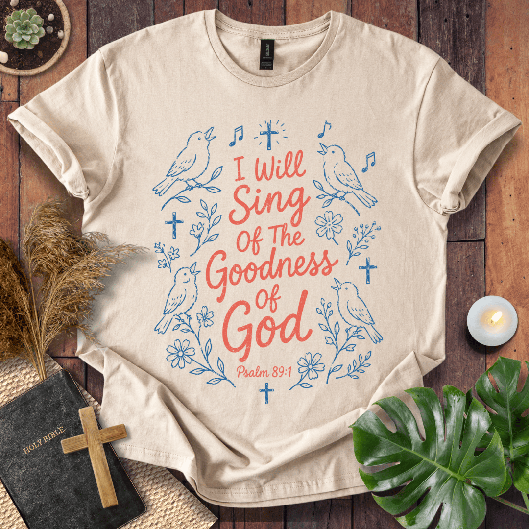 A beige short-sleeve graphic T-shirt laid flat on a rustic wooden surface, surrounded by decorative props including a small cross, a Bible, dried grass, a candle, and green leaves. The shirt has a blue and coral-red printed design with birds, flowers, musical notes, and the text 'I Will Sing Of The Goodness Of God Psalm 89:1'.