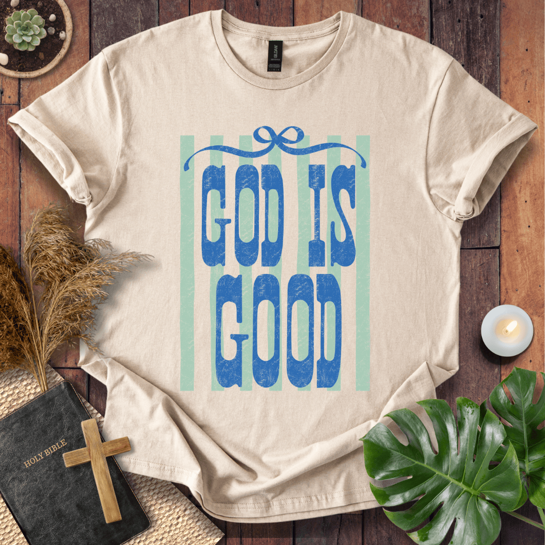 A beige short-sleeve graphic T-shirt laid flat on a rustic wooden surface, featuring the words 'GOD IS GOOD' in blue letters over pale mint-green vertical stripes. The shirt is styled with a small cross, a Bible, dried grass, a plant leaf, and a candle.