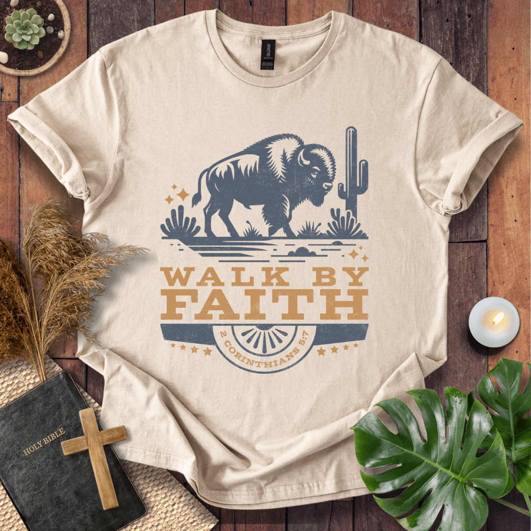 Beige t-shirt with a graphic of a bison and cactus, featuring the text 'Walk by Faith', on a wooden surface with decorative items.