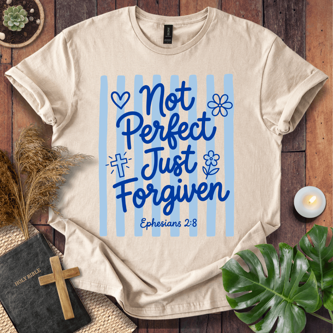 A beige graphic t-shirt laid flat on a rustic wooden surface, surrounded by decorative props including a Bible, a wooden cross, dried grass, a small candle, and green leaves. The t-shirt features a blue graphic with the text 'Not Perfect Just Forgiven Ephesians 2:8' on a background of light blue vertical stripes.