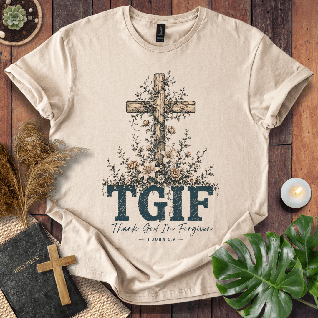 A beige graphic t-shirt laid flat on a rustic wooden surface, featuring a floral cross design and the text 'TGIF Thank God I'm Forgiven — 1 John 1:9'. The shirt is styled with a small wooden cross, a Bible, dried grass, a candle, and green leaves.