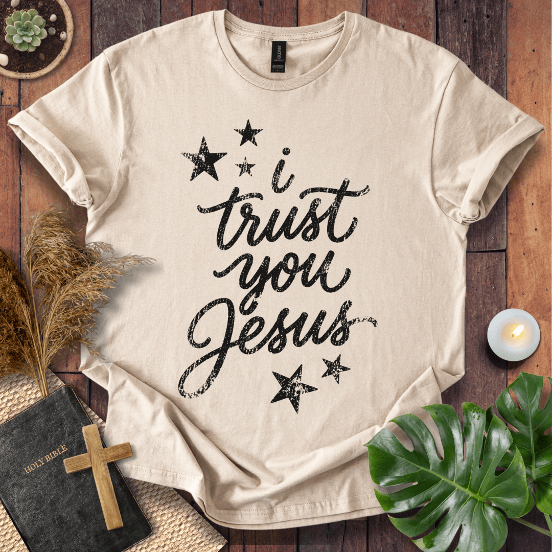 A beige short-sleeve T-shirt laid flat on a rustic wooden surface, printed with the phrase 'i trust you Jesus' in black script and surrounded by black stars. The shirt is styled with a Bible, wooden cross, dried grass, a small candle, and green leaves.