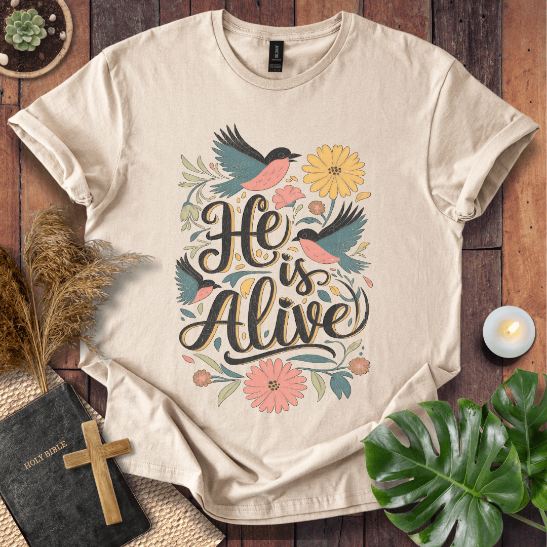 He Is Alive" Christian Easter t shirt for women with birds and flowers on a natural cream shirt, styled on a rustic wood background.