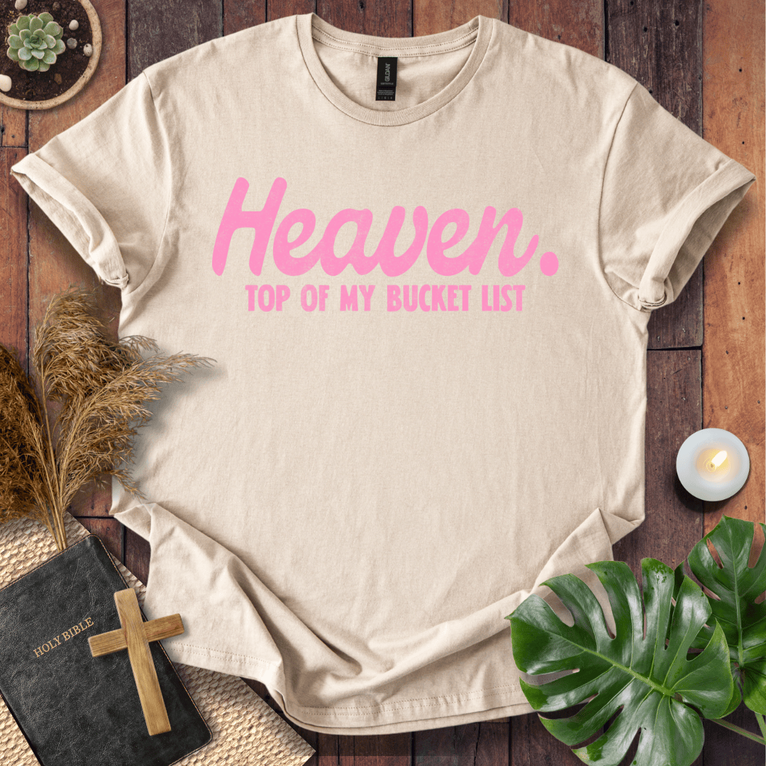 A beige short-sleeve graphic T-shirt laid flat on a rustic wooden surface, with pink text reading 'Heaven. TOP OF MY BUCKET LIST.