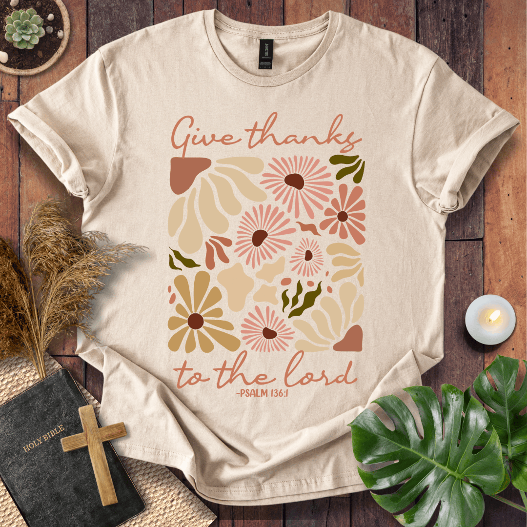 Give Thanks to the Lord Psalm 136:1 white graphic t-shirt with retro boho rust and sage wildflowers on a wood background.