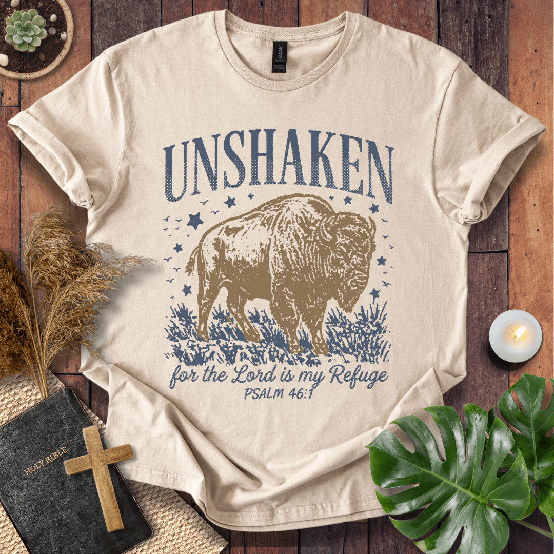A beige graphic t-shirt featuring a brown bison and blue text reading 'UNSHAKEN for the Lord is my Refuge PSALM 46:1', styled in a flat-lay on a wooden background with a book, cross, pampas grass, and green leaves.