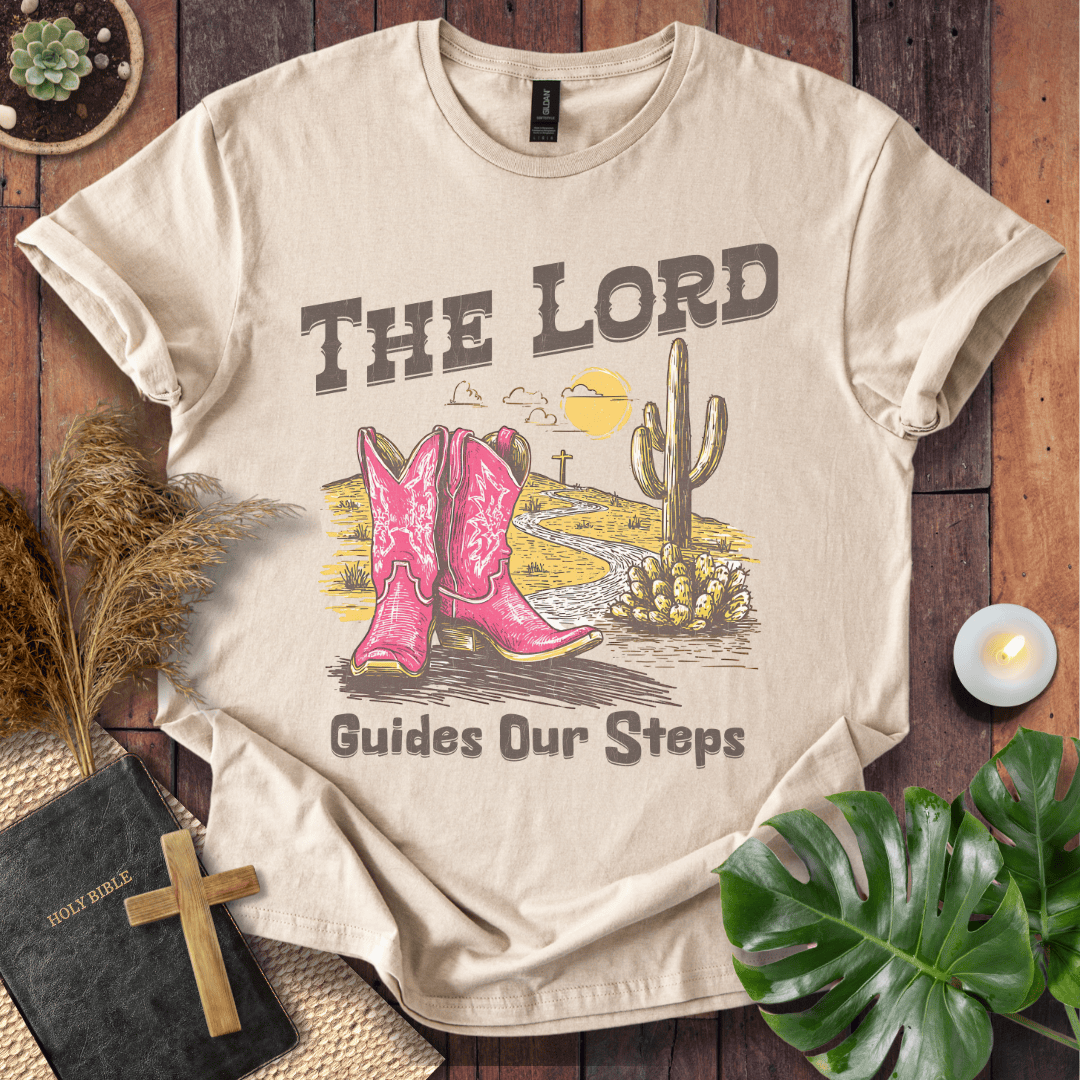 t-shirt with pink cowboy boots and "The Lord Guides Our Steps" text in a desert landscape with a cross and cactus.