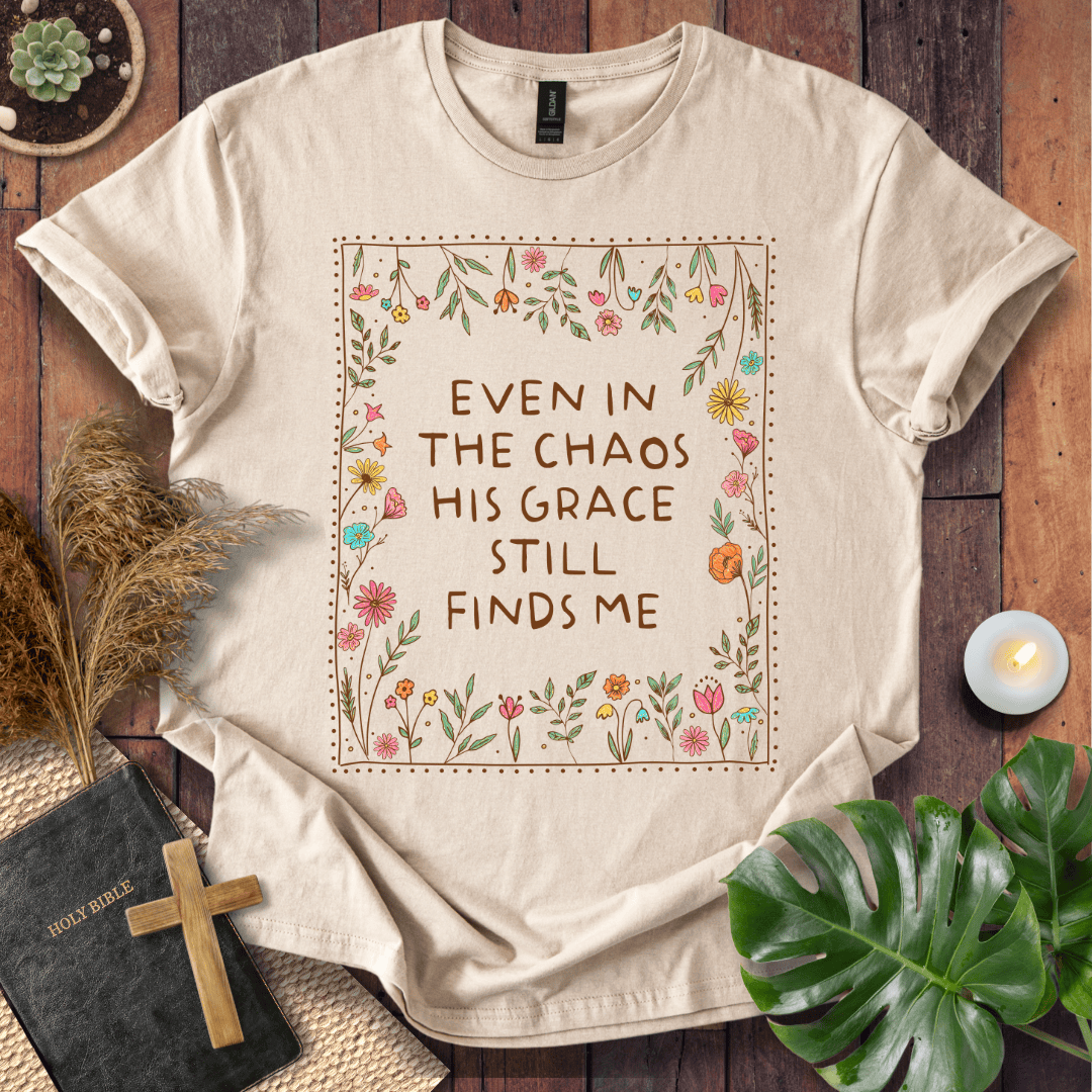 A beige graphic t-shirt laid flat on a rustic wooden background, surrounded by decorative props including a Bible, a wooden cross, dried grass, a small candle, and green leaves. The t-shirt features a central square floral border with the brown text 'EVEN IN THE CHAOS HIS GRACE STILL FINDS ME'.