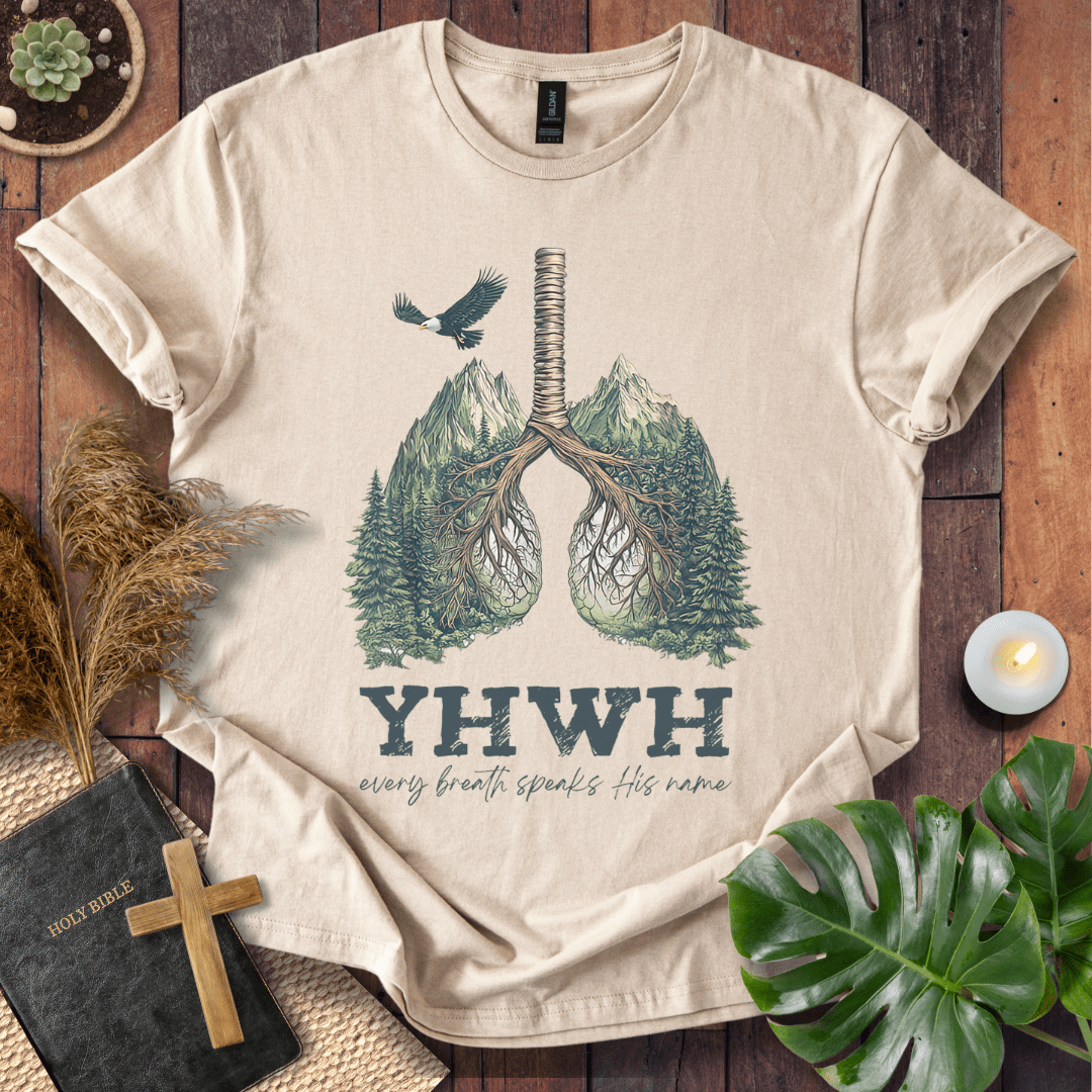 Christian t-shirt with YHWH design, tree-lung illustration, mountains, and “Every Breath Speaks His Name” text, laid flat with Bible and plants.